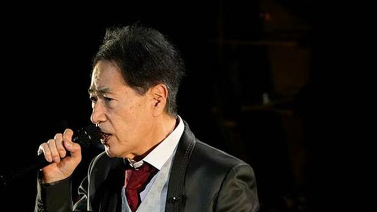 赤いネクタイ姿でマイクを持つ稲垣潤一