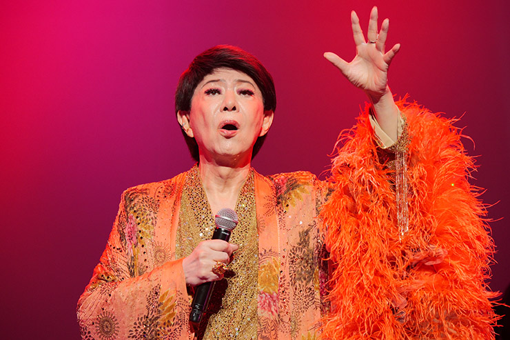 橙色の衣装でマイクを持ち、左手を上げる美川憲一