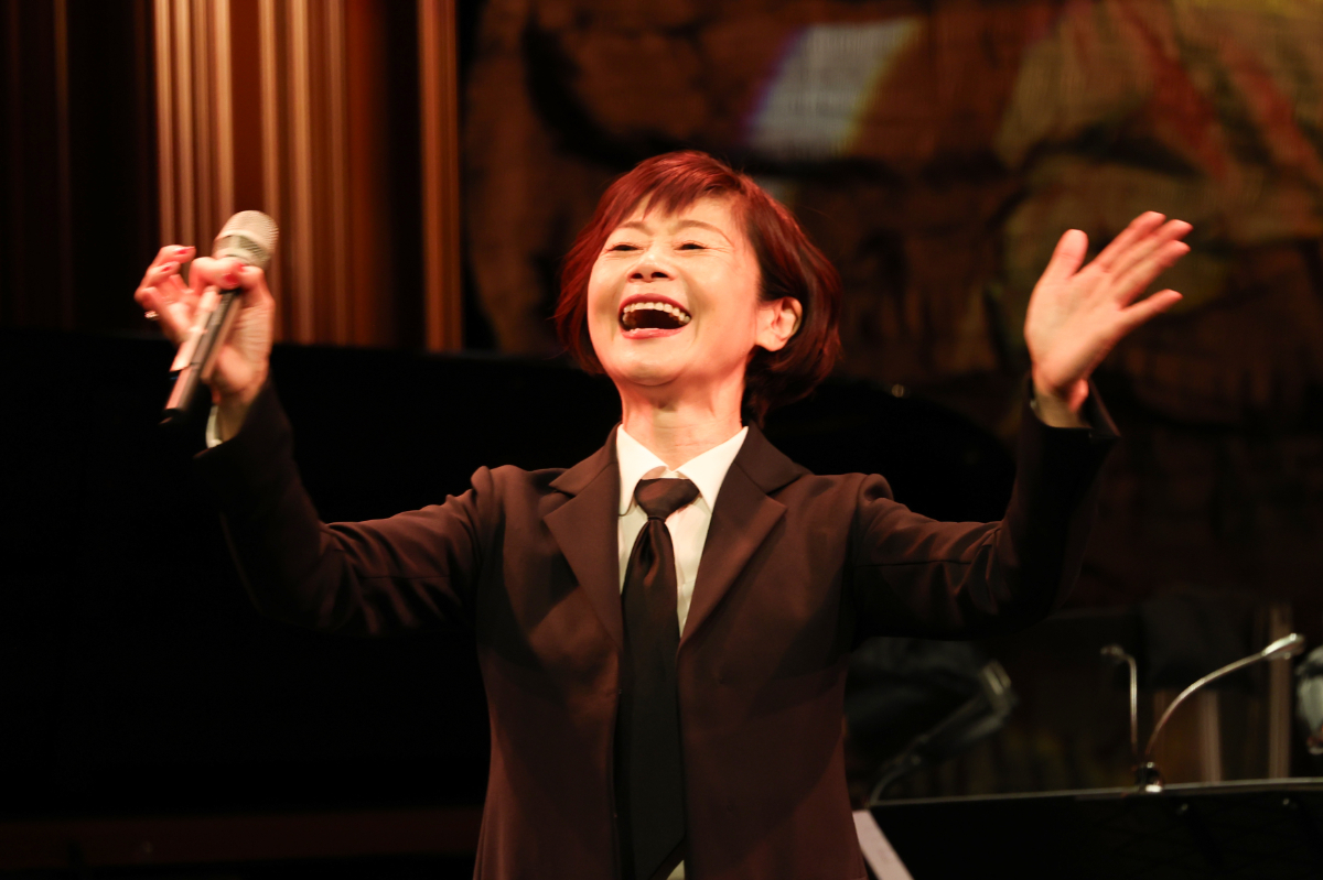 黒いスーツとネクタイ姿で両腕を広げ、マイクを手に歌う神野美伽